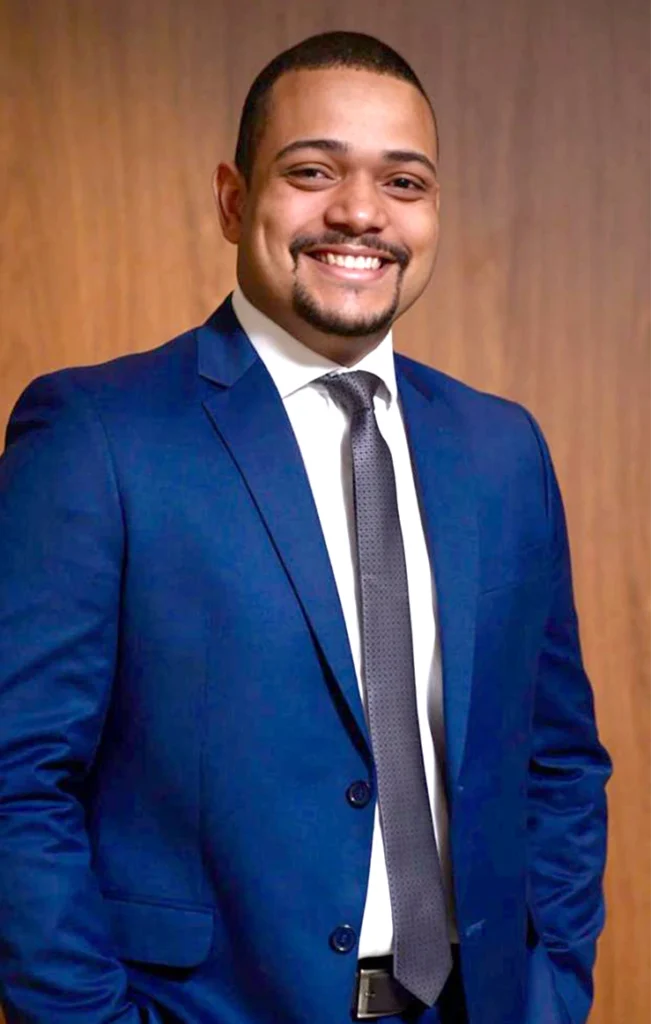 Foto do advogado Bruno da Silva Adelino , atuante nas áreas de Processo Civil e Direito Público. Usando um terno azul e gravata grafite na frente de um fundo de madeira.